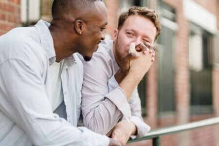 Two men smiling and holding hands while leaning on a railing outdoors.