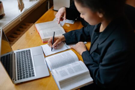 Two students engaged in study, taking notes with books and a laptop on a wooden desk indoors.