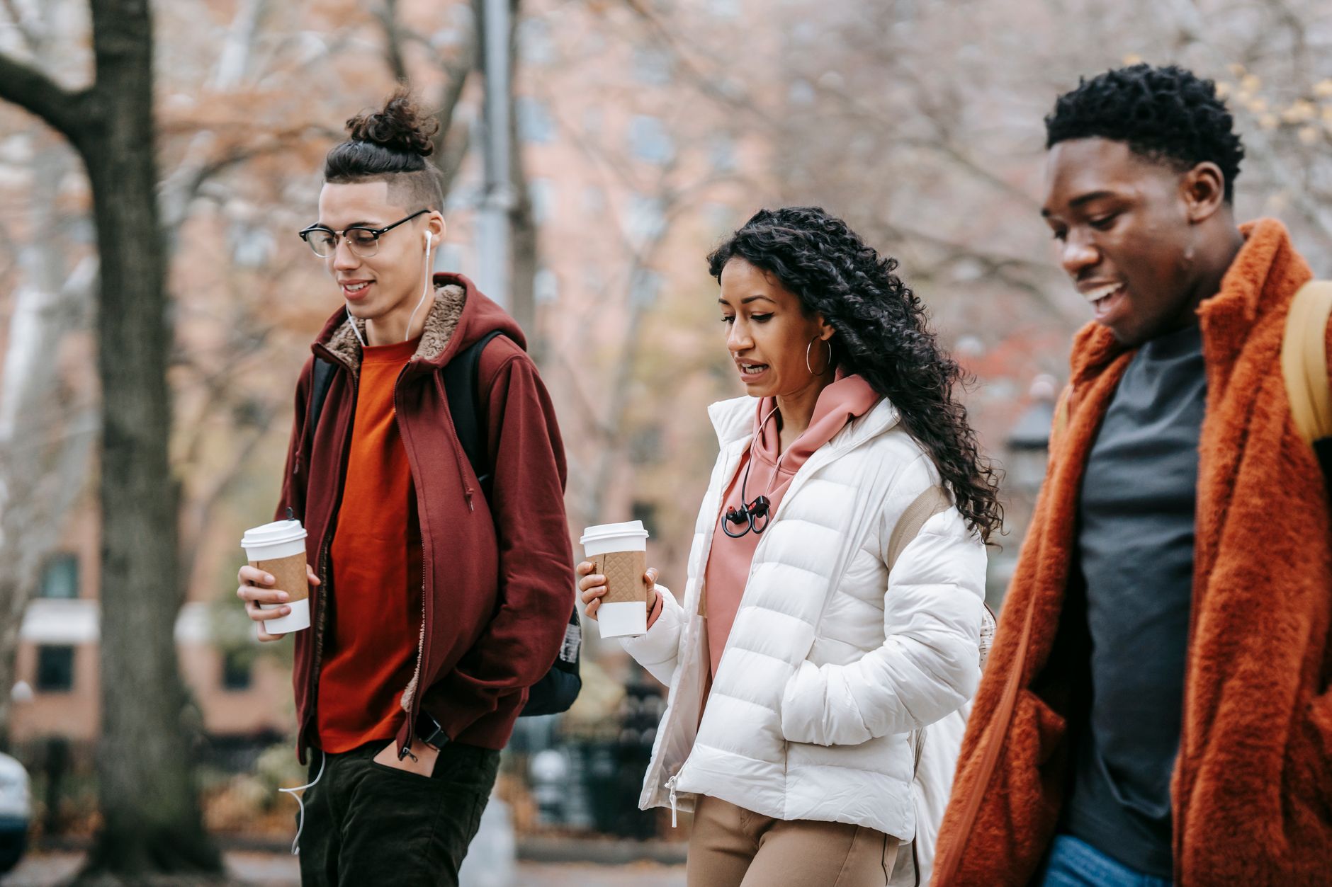 Jeunes amis multiraciaux confiants en tenue d'extérieur et sacs à dos discutant tout en se promenant dans une rue de la ville près d'arbres avec des gobelets en papier avec des boissons et des écouteurs en journée dans un parc