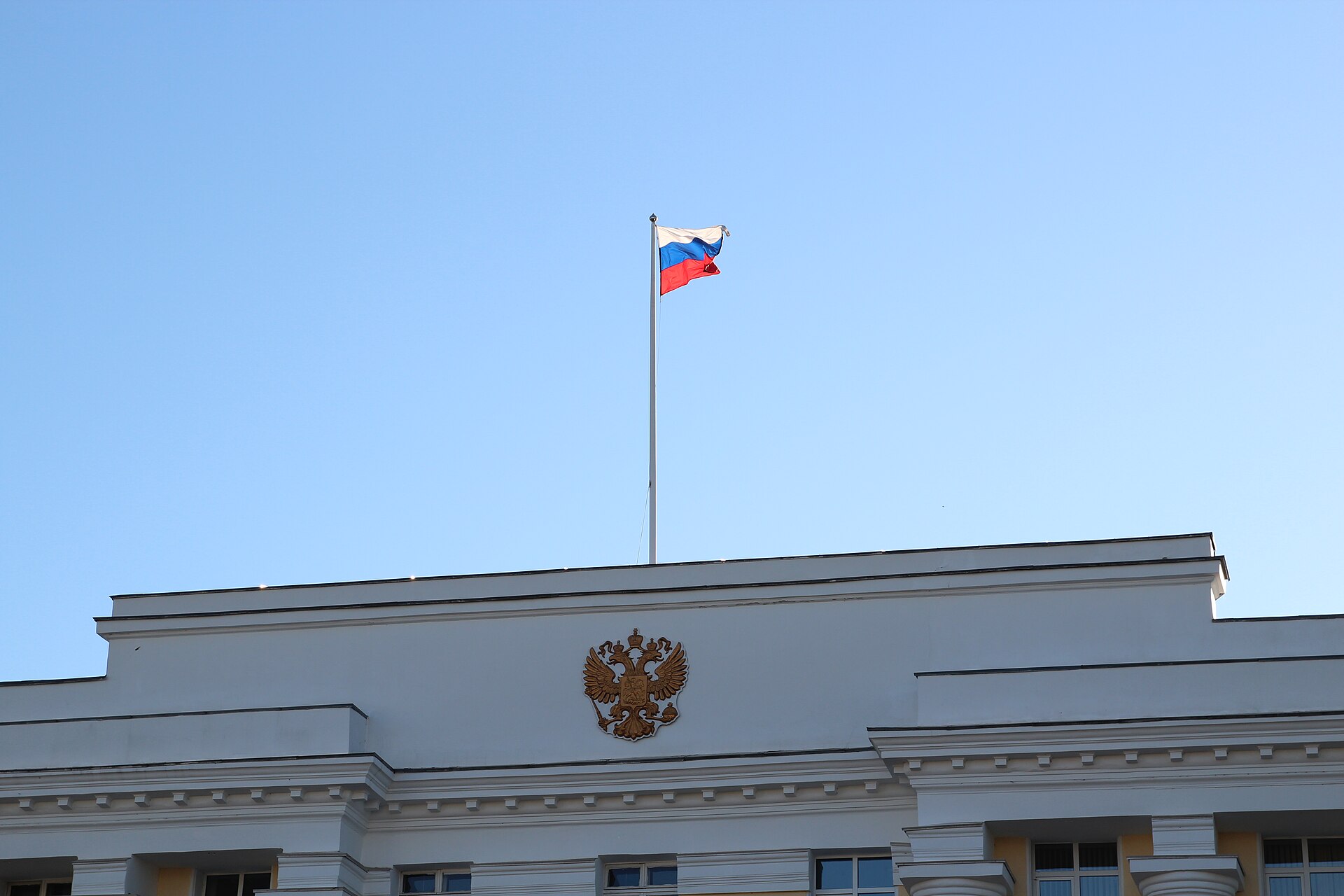 Drapeau tricolore blanc, bleu et rouge flottant au-dessus d’une façade officielle ornée d’un aigle bicéphale