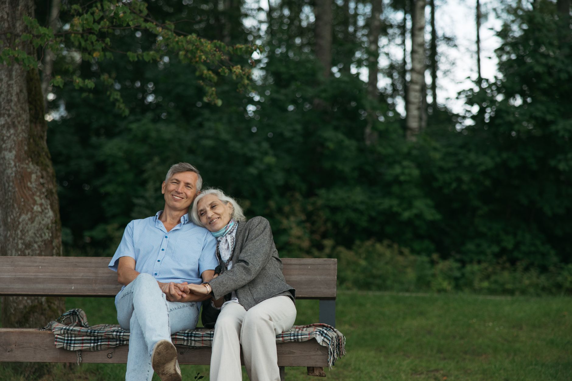 Un couple de seniors souriants, main dans la main, assis sur un banc en bois dans un parc, devant des arbres.