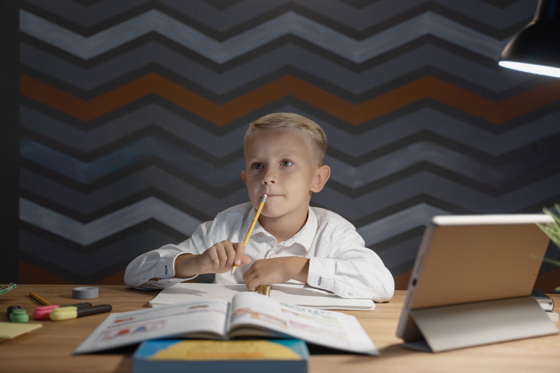 Un garçon en chemise blanche mordille un crayon à son bureau, devant un livre ouvert, un cahier et une tablette, sous une lampe.