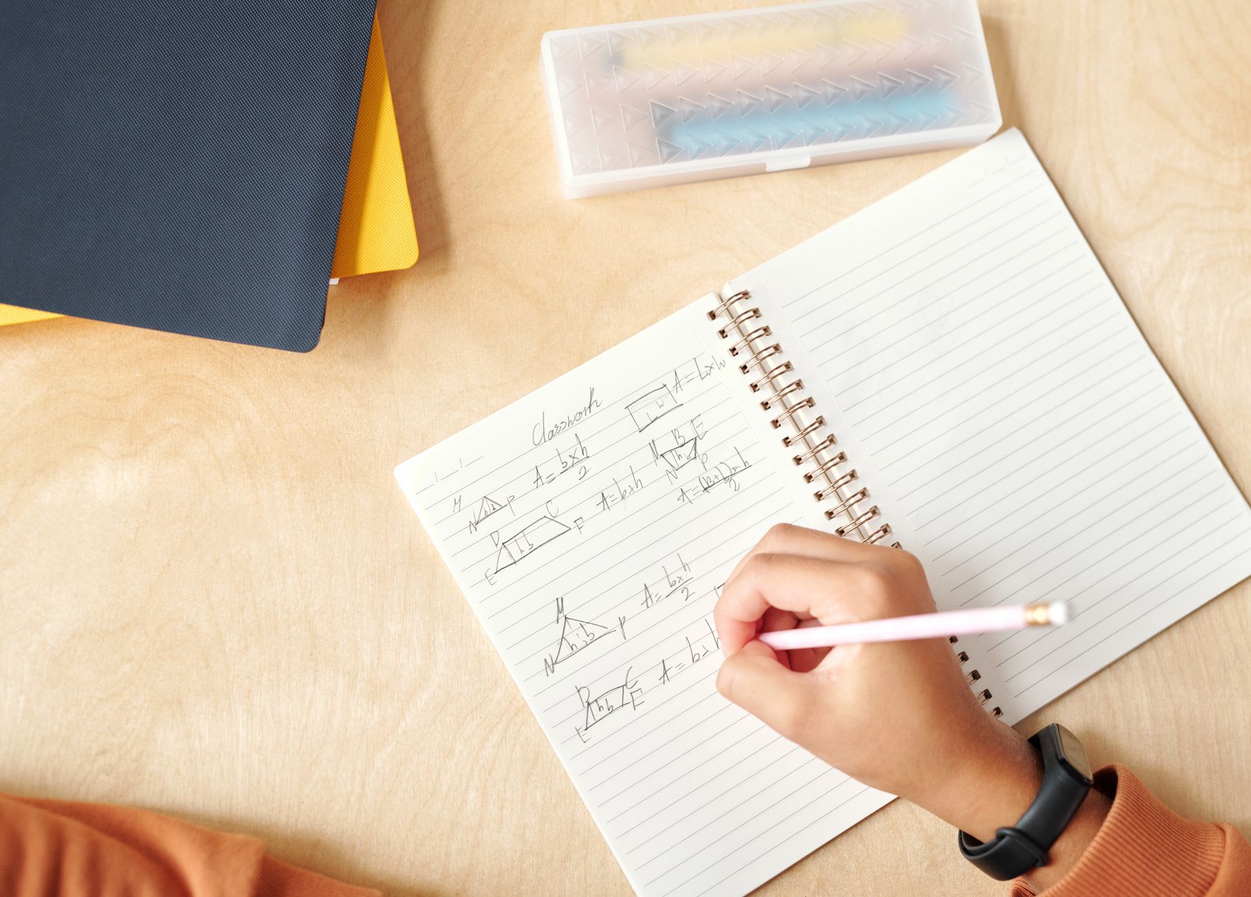 Vue de dessus d’une main qui écrit au crayon dans un cahier à spirale, avec schémas et formules de géométrie, sur un bureau en bois clair.